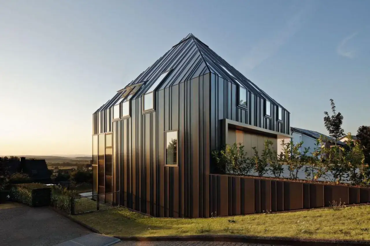 Nowoczesny dom z ciemną blachą na rąbek elewacja, z licznymi oknami i zielenią.