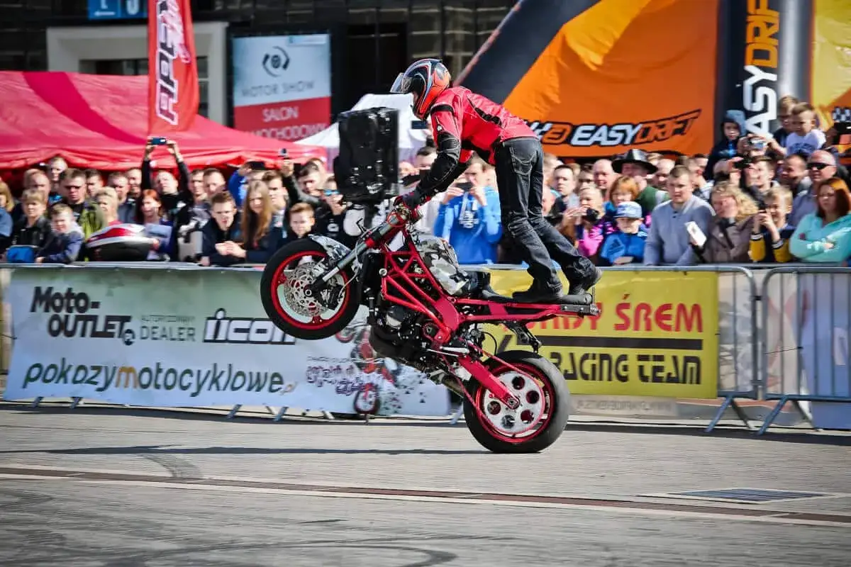 Motocykl do stuntu na tylnym kole, kierowca w czerwonej kurtce, tłum widz&oacute;w za barierkami.