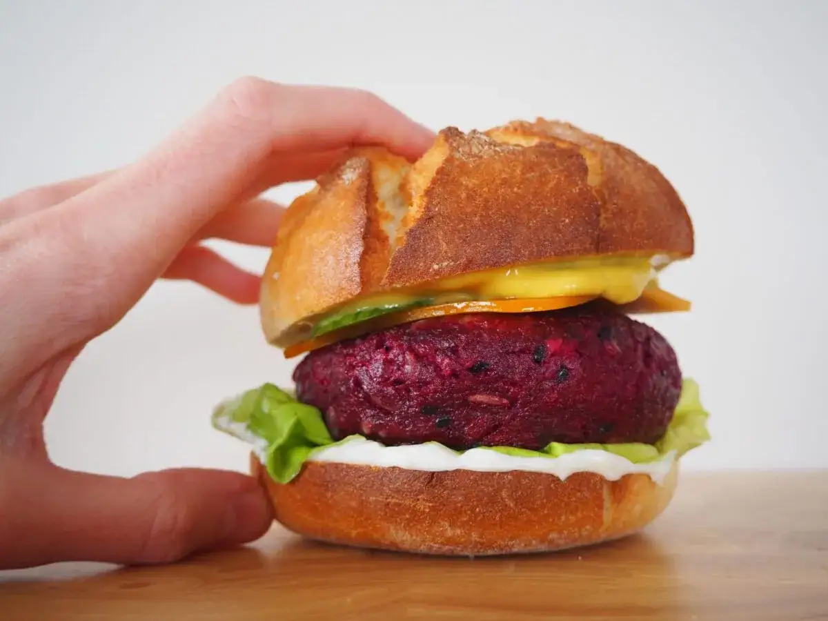 Ręka trzyma apetyczny burger z buraka, z chrupiącą bułką, sałatą i sosem.