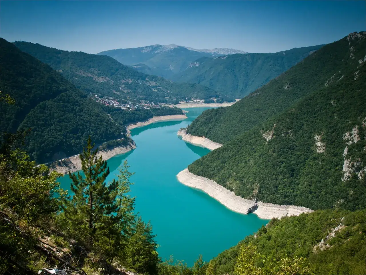 Turkusowe Pivsko Jezero otoczone zielonymi g&oacute;rami, z widocznym miasteczkiem na brzegu.
