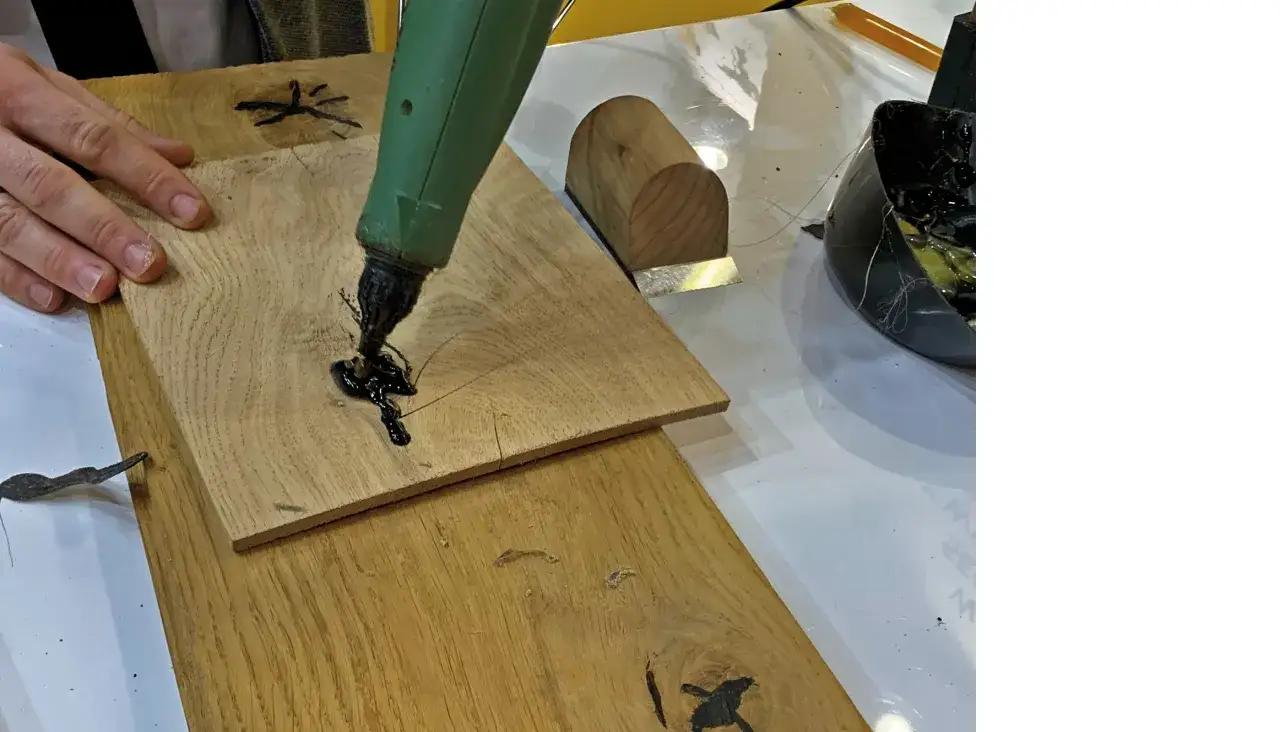 Ręka aplikuje czarną masę z pistoletu, by czym uzupełnić ubytki w drewnie.