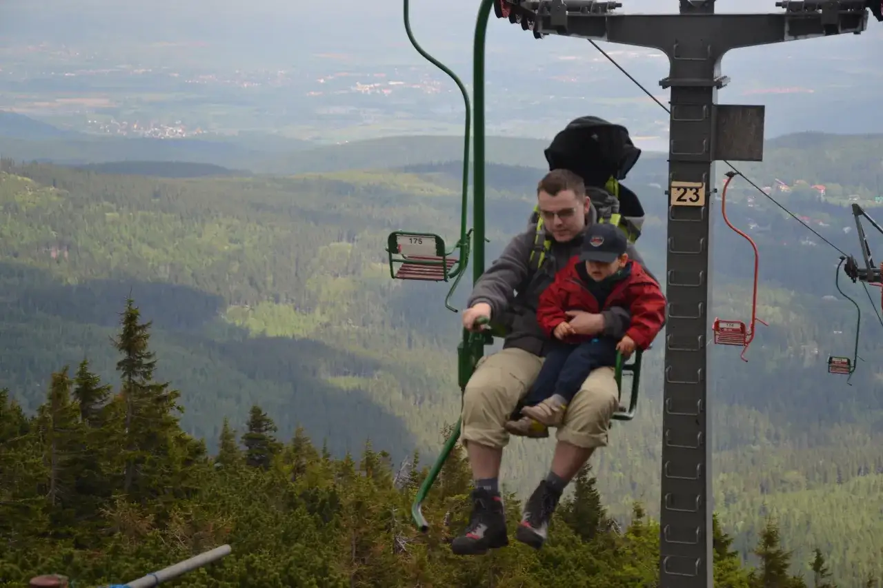 Ojciec z dzieckiem na wyciągu krzesełkowym, podziwiający widok na gęsty las. Zastanawiają się, kt&oacute;ry szlak na Śnieżkę najłatwiejszy.