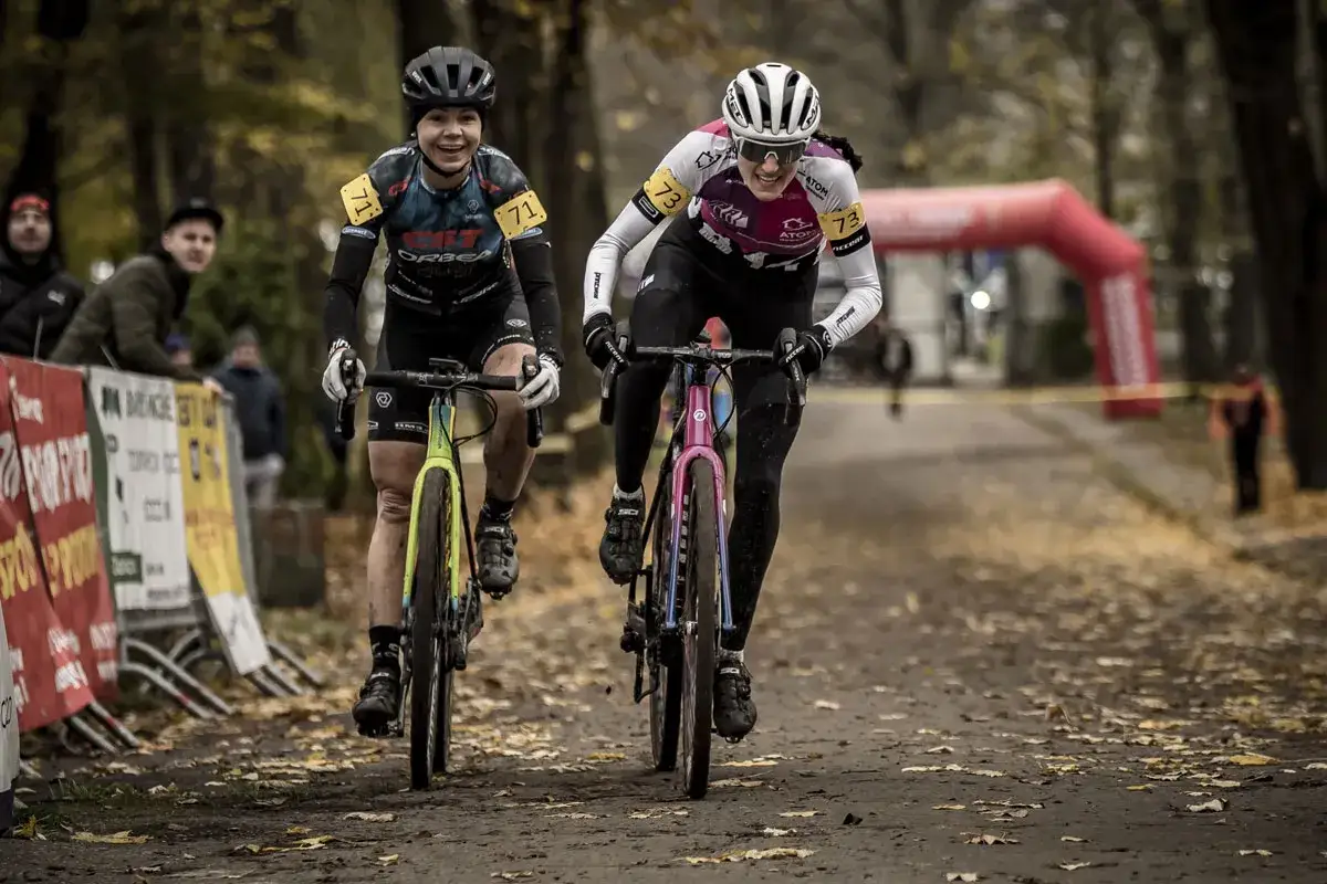 Dwie kolarki ścigają się na rowerach gravelowych. Jedna z nich jedzie na rowerze road plus w kolorze ż&oacute;łto-zielonym, druga na r&oacute;żowo-niebieskim.