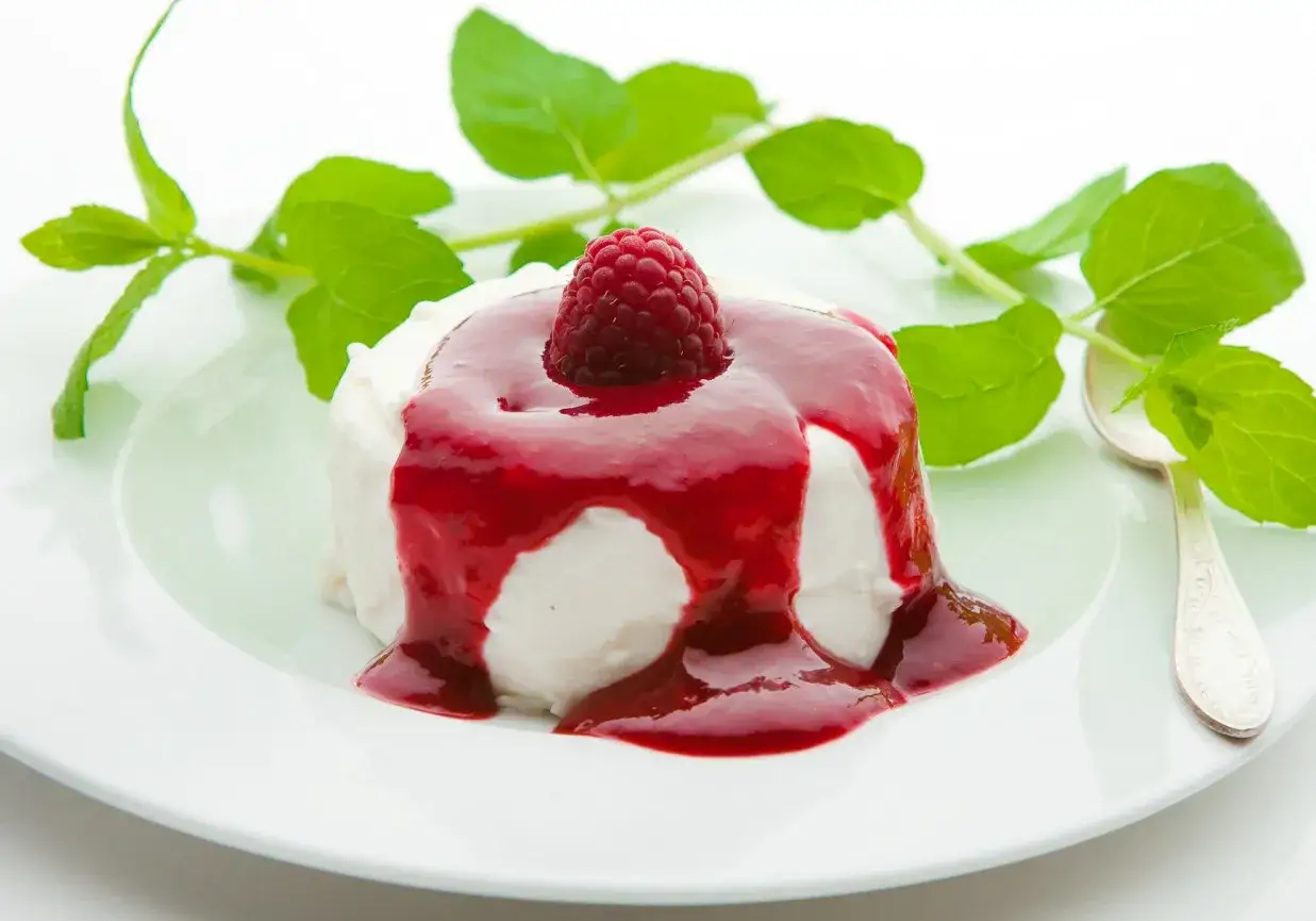 Panna cotta na śmietance, polana sosem malinowym i ozdobiona maliną. Obok listki mięty i łyżeczka.