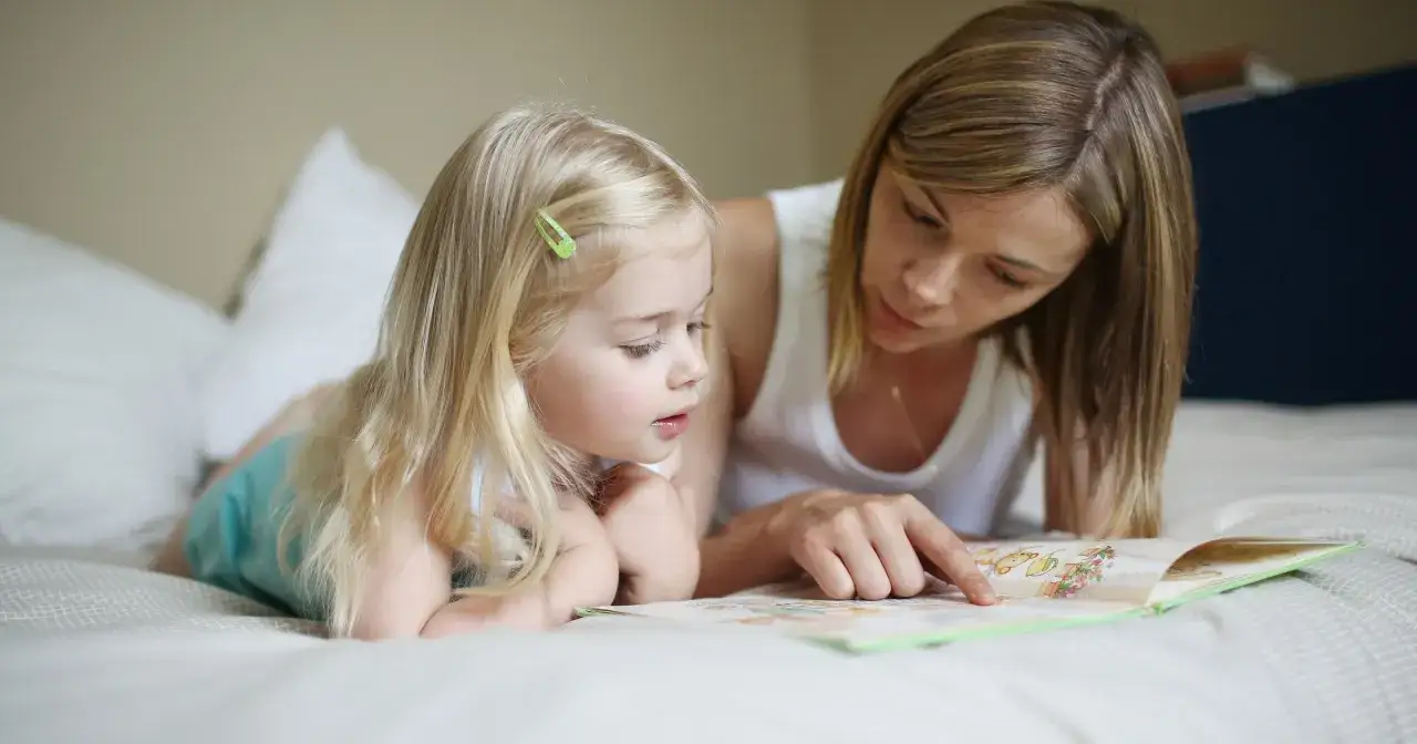 Mama uczy córkę czytać metodą sylabową, wskazując palcem na ilustracje w książce.