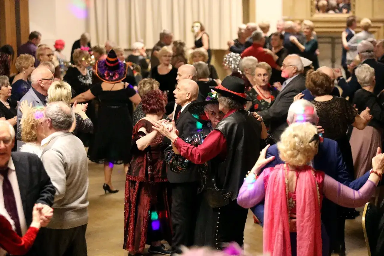 Wesoła potańcówka dla seniorów w Warszawie. Ludzie w różnym wieku bawią się w rytm muzyki, niektórzy w strojach.