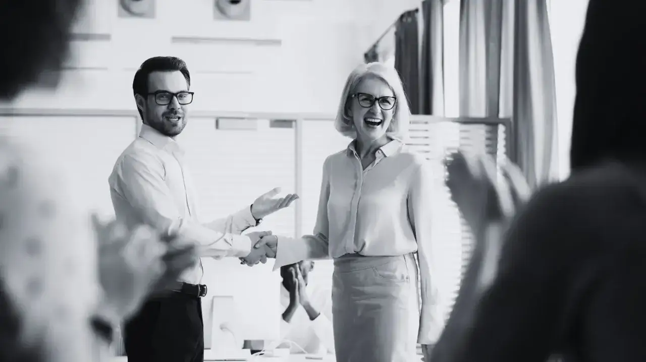 Mężczyzna i kobieta ściskają sobie dłonie, otoczeni ludźmi klaszczącymi. Widać uśmiechniętą twarz kobiety, kt&oacute;ra otrzymuje mądre komplementy.