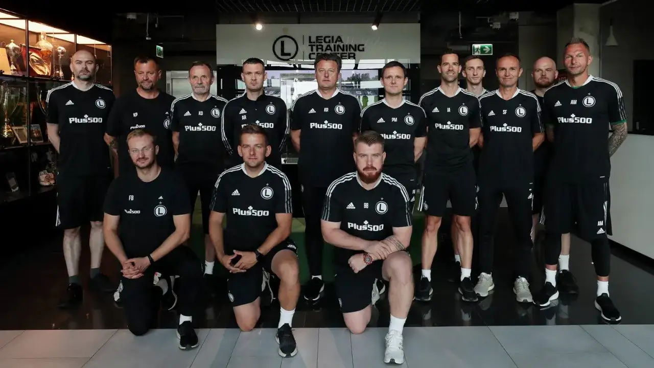 Grupa zawodników Legii Warszawa w czarnych strojach treningowych pozuje do zdjęcia w centrum treningowym.