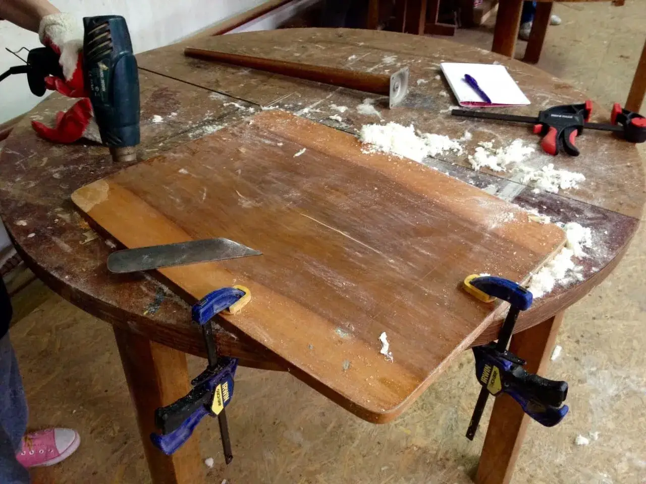 Praca nad tym, jak usunąć lakier z drewna. Na stole leży opalarka, skrobak i ściski.