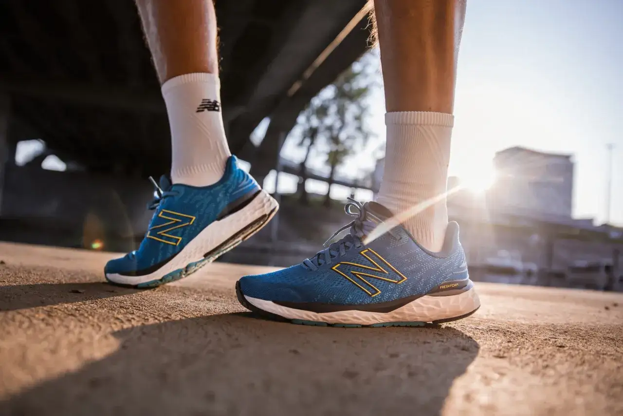 Biegacz w białych skarpetkach i niebieskich butach do biegania naturalnego z ż&oacute;łtym logo N. Słońce przebija się przez miejską zabudowę.