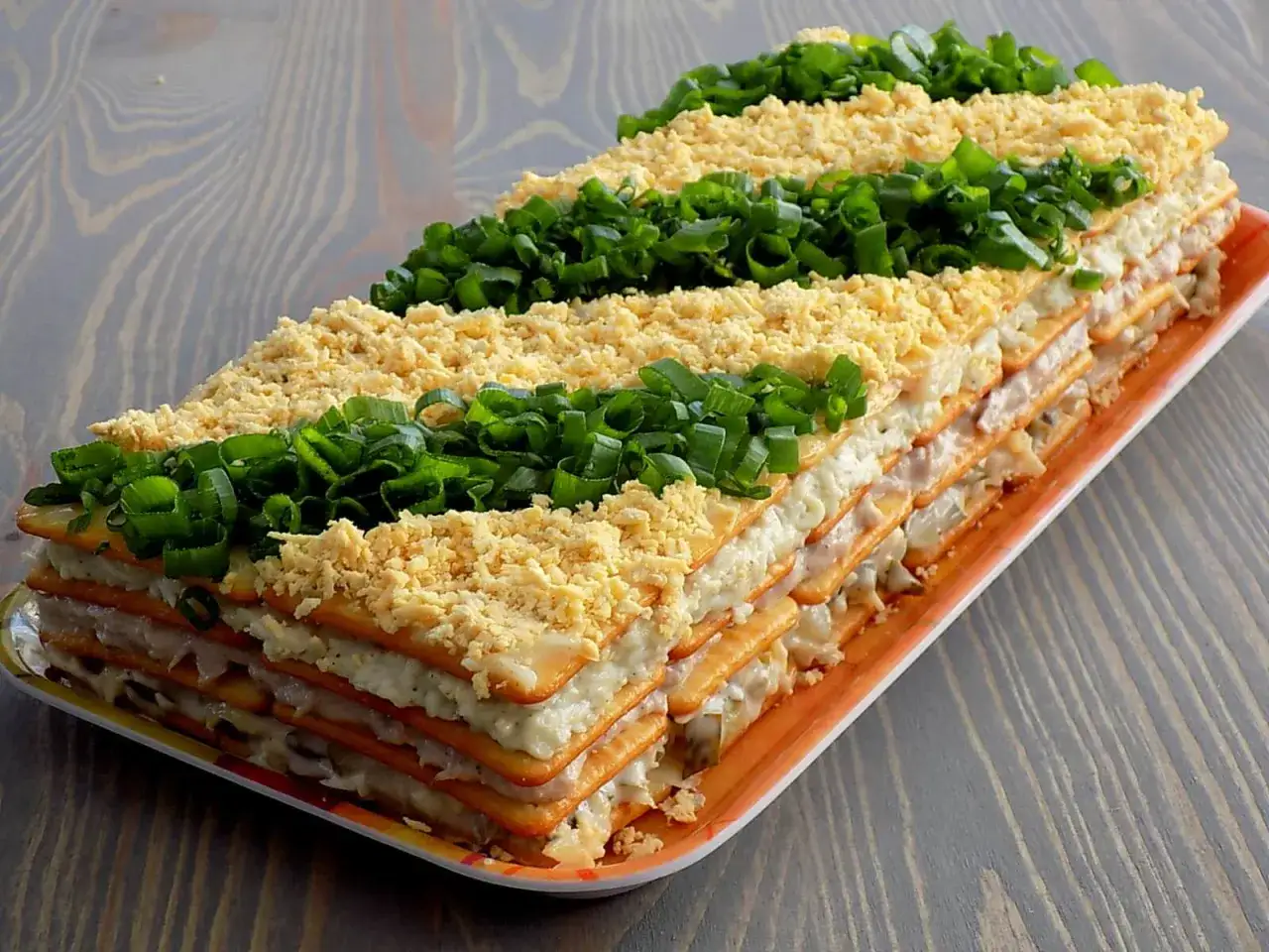 Warstwowa sałatka na krakersach z jajkiem i szczypiorkiem, ułożona na paterze.