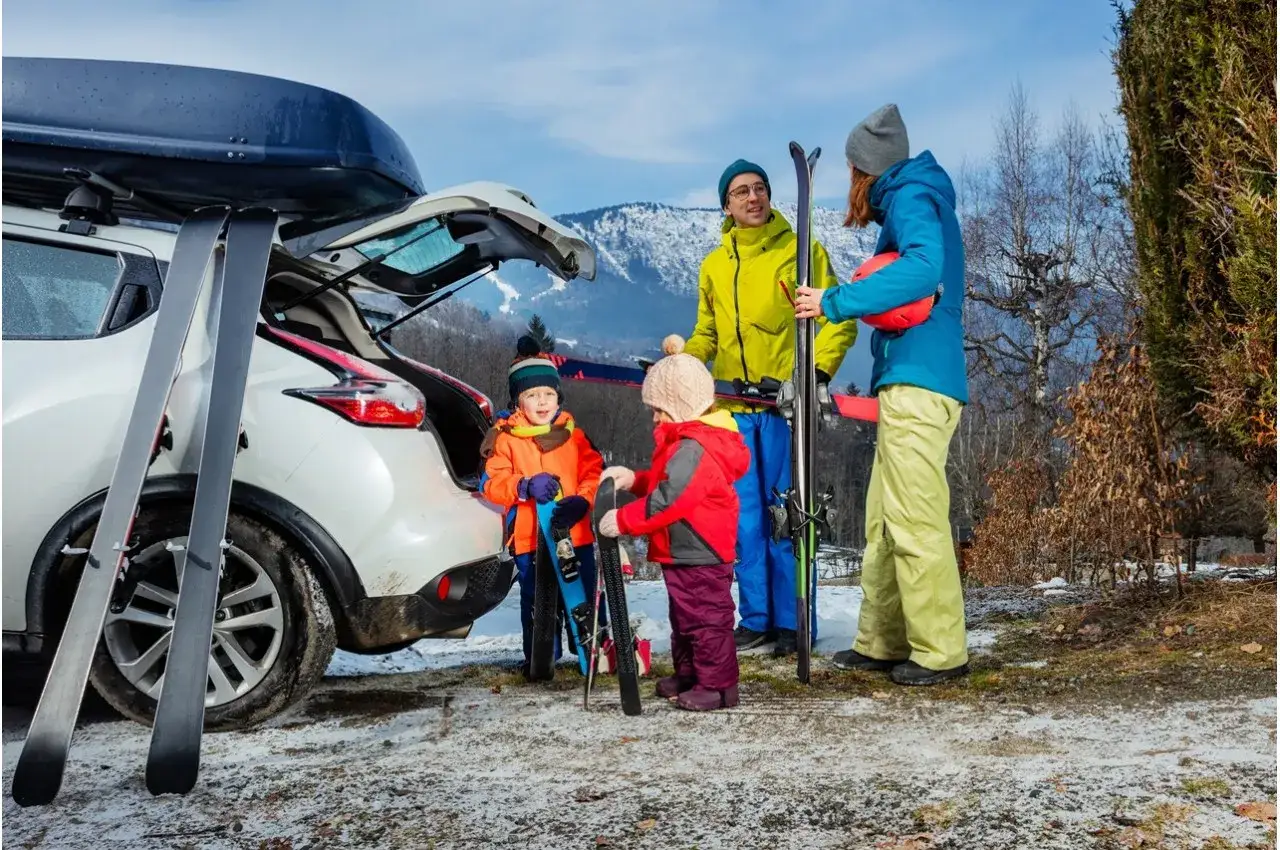 Rodzina z nartami przy samochodzie z boxem dachowym, gotowa na zimową przygodę.