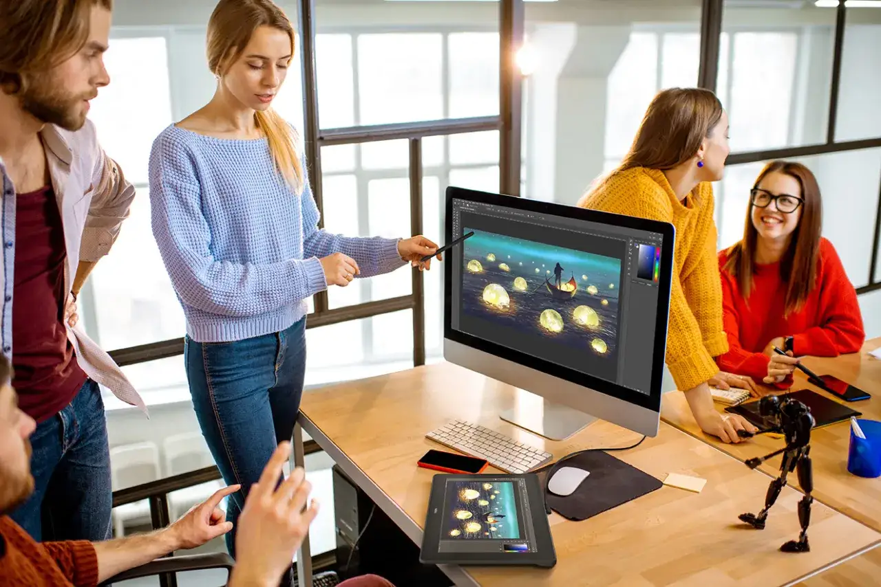 Zesp&oacute;ł pracuje nad projektem graficznym, korzystając z **aplikacji do rysowania na tablecie**. Na ekranie komputera widać magiczną scenę z łodzią i świetlistymi kulami.