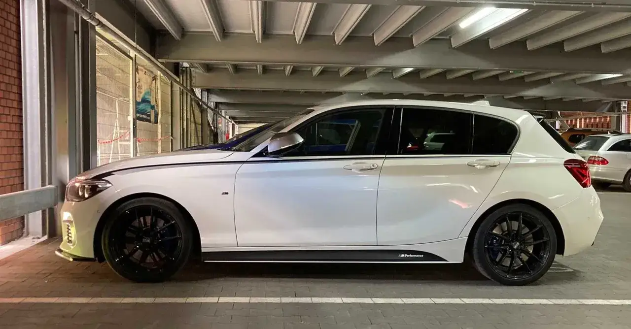 Białe BMW 140i z czarnymi felgami i naklejkami M Performance, zaparkowane w garażu.