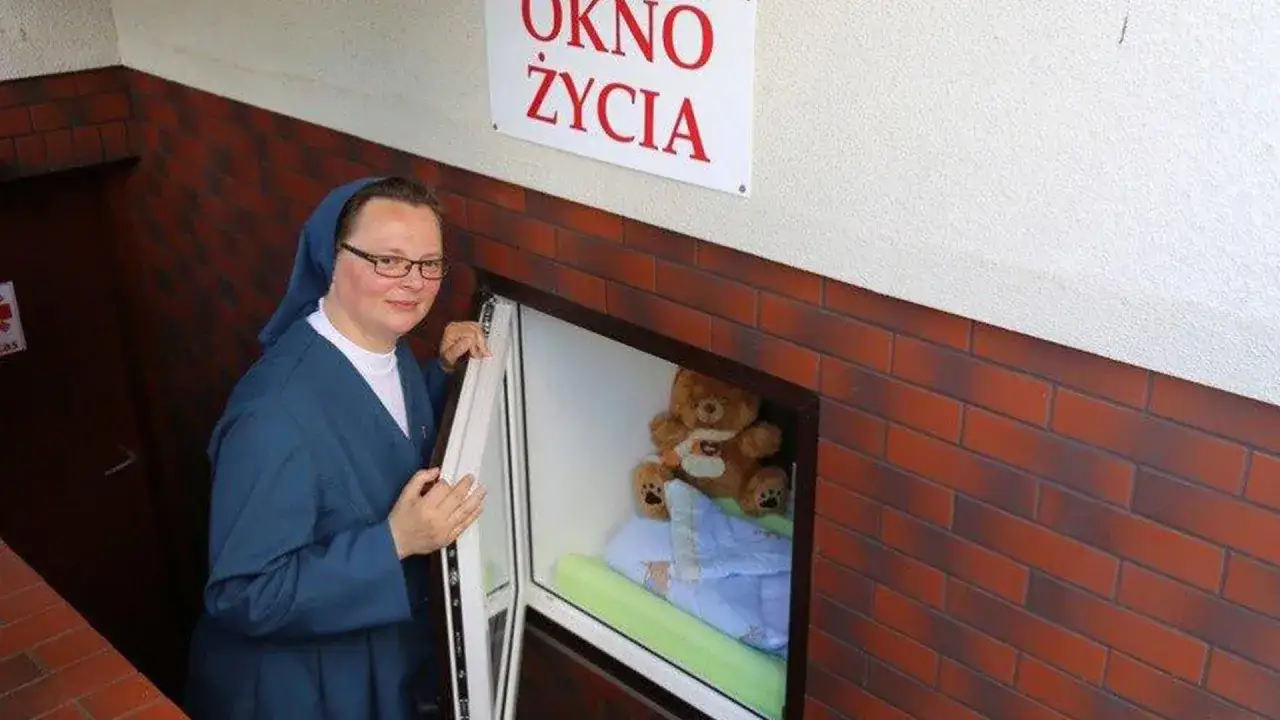 Siostra otwiera okno życia, w którym czeka pluszowy miś i kołderka.
