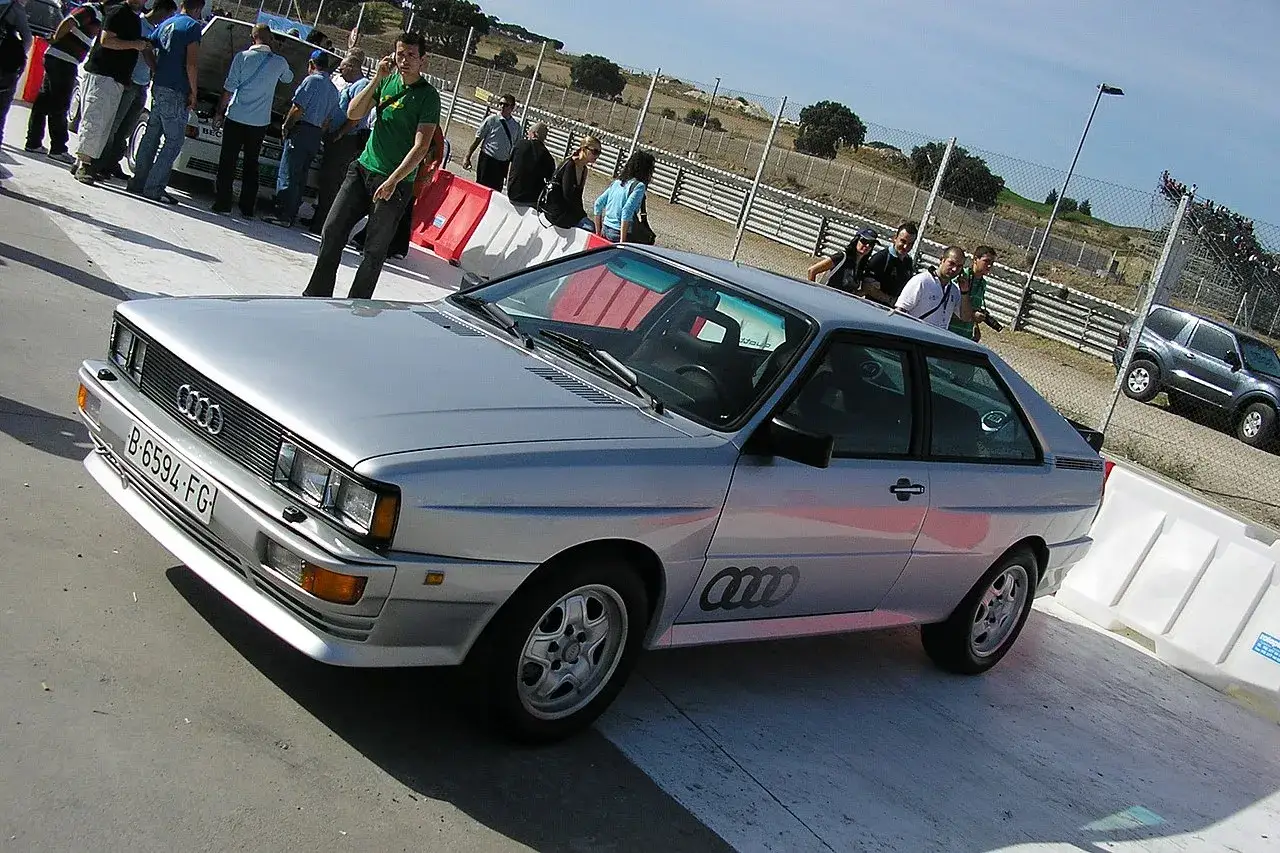 Srebrne audi quattro z lat 80. na torze wyścigowym, otoczone przez widzów i bariery.