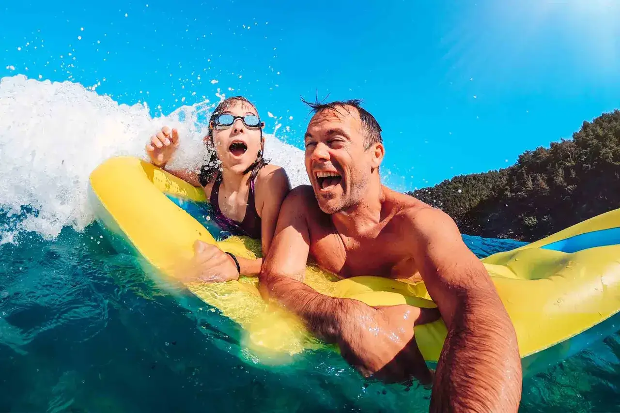 Ojciec i córka śmieją się na żółtym materacu do pływania, gdy fala rozbija się obok nich na turkusowym morzu.