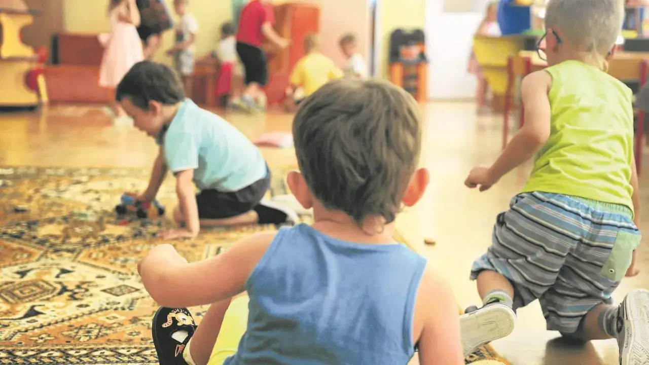 Dzieci bawią się na dywanie. Jedno z nich, z tyłu, ma niebieską koszulkę. W tle widać inne dzieci. Obrazek może ilustrować zapalenie oskrzeli u dzieci, gdy choroba nie przeszkadza w zabawie.