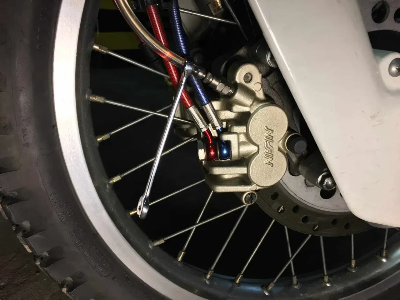 Klucz nasadowy przy zacisku hamulcowym motocykla, przygotowanie do wymiany płynu hamulcowego. Widać przewody hamulcowe i tarczę.