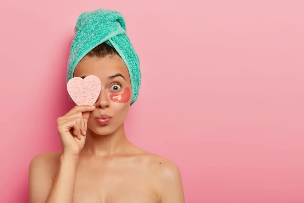 Krok po kroku pielęgnacja twarzy: dziewczyna z ręcznikiem na głowie, płatkami pod oczy i gąbką w kształcie serca.
