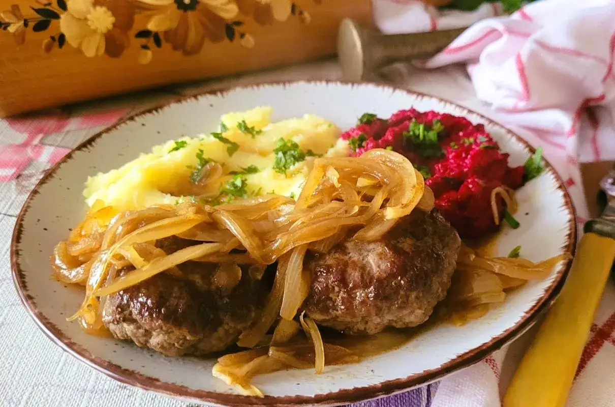 Dwa soczyste rumsztyki, polane duszonymi cebulkami, podane z purée ziemniaczanym i buraczkami. Pyszny bryzol na talerzu.