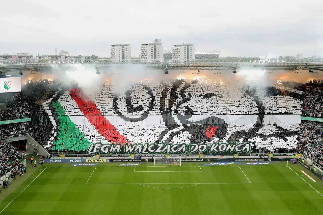 Trybuna stadionu wypełniona kibicami tworzącymi transparent z hasłem 