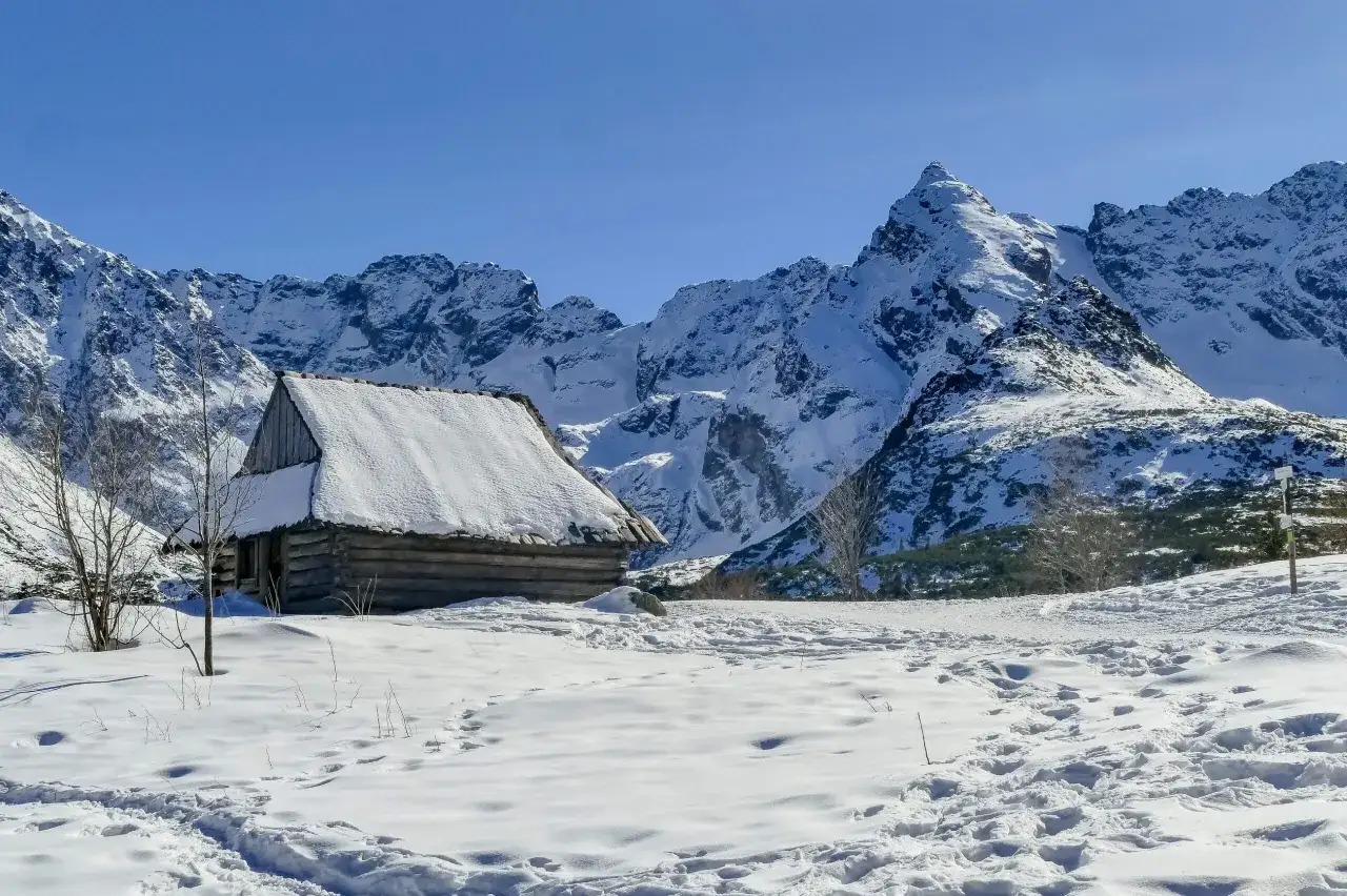 Drewniana chata pokryta śniegiem na tle ośnieżonych gór. Ślady na śniegu wskazują szlak na Nosal.