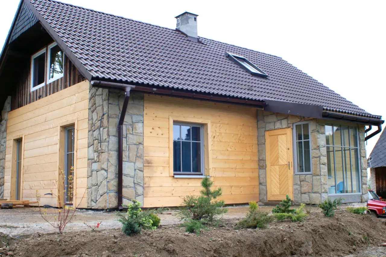 Nowy dom z piaskowca i drewna, z brązowym dachem, oknami i drzwiami. Wok&oacute;ł młode drzewka i ziemia.