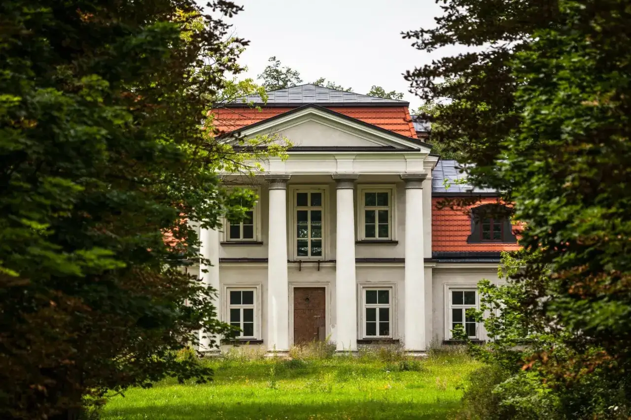 Klasyczny dworek z kolumnami i czerwoną dachówką, otoczony zielenią. Idealne miejsce dla szukających dworki na sprzedaż.