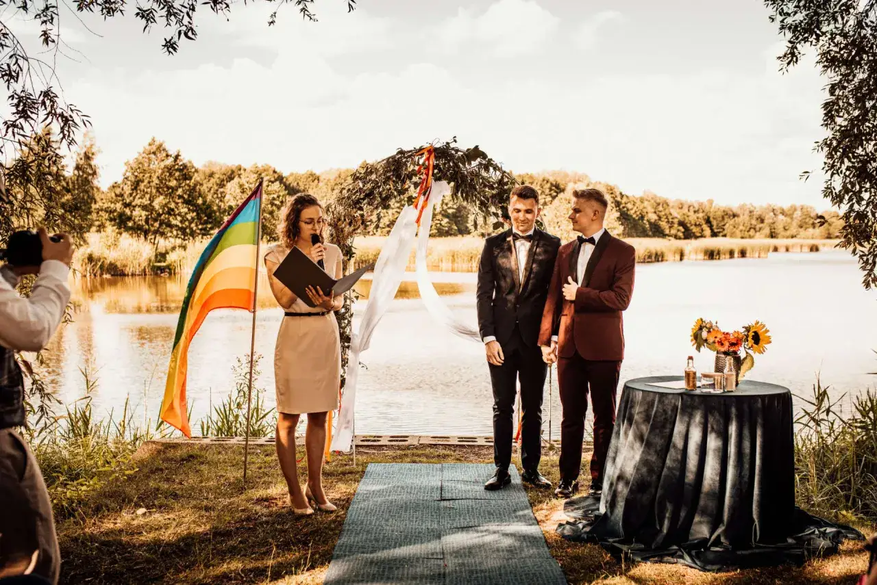 Uroczystość nad jeziorem, gdzie można wziąć ślub homoseksualny. Dwóch panów młodych, tęczowa flaga i urzędniczka udzielająca ślubu.