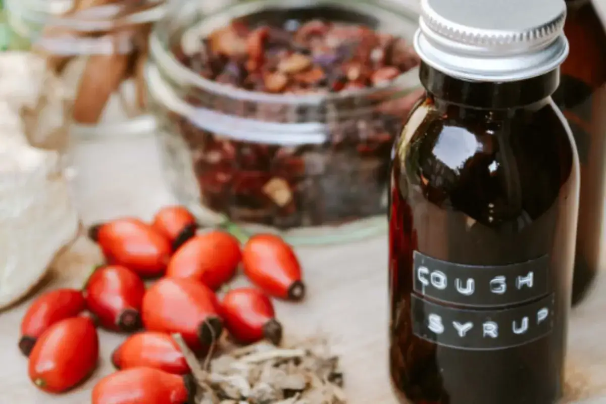 Naturalne składniki, jak dzika r&oacute;ża i suszone zioła, do przygotowania syropu na kaszel. W butelce ciemny płyn z etykietą 