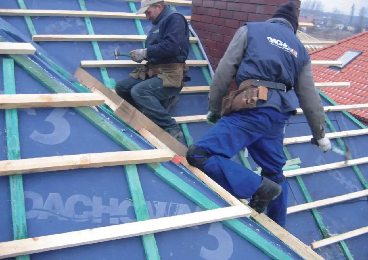 Dw&oacute;ch budowlańc&oacute;w pracuje nad dachem, montując membranę dachową. Widać drewniane łaty i elementy konstrukcyjne.
