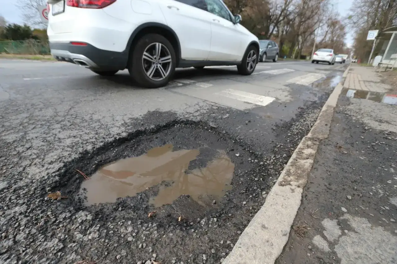 Ogromna poprzeczna nierówność jezdni z wodą, zagrażająca samochodom. Biały SUV zbliża się do dziury.