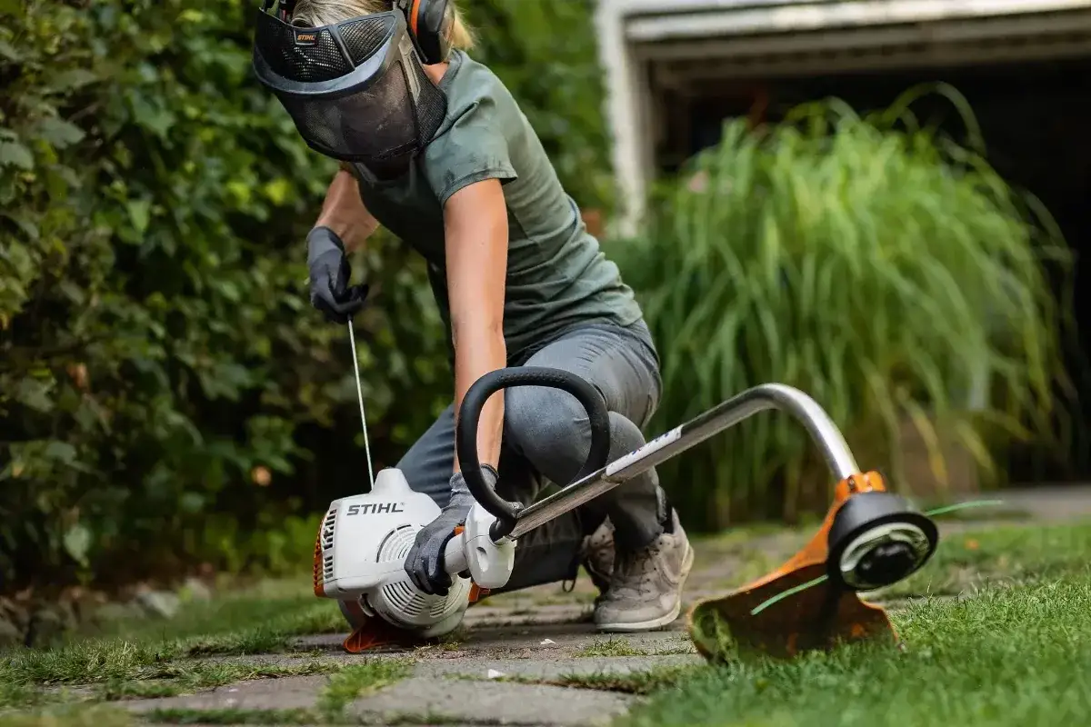 Osoba w ochronnej masce i słuchawkach przygotowuje się do pracy, chwytając za linkę rozruchową kosy spalinowej.