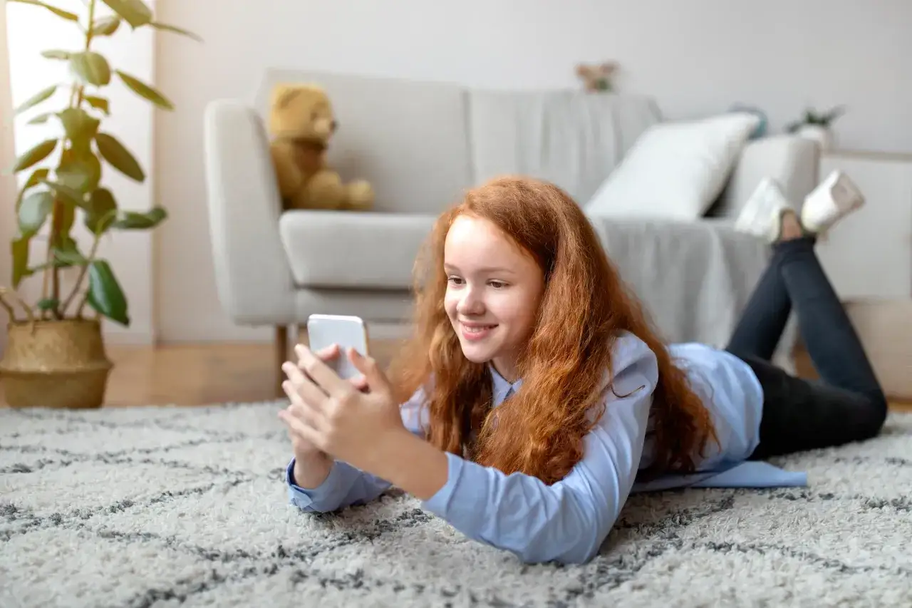 Uśmiechnięta dziewczynka z rudymi włosami leży na dywanie, trzymając w dłoniach sw&oacute;j pierwszy telefon dla dziecka.