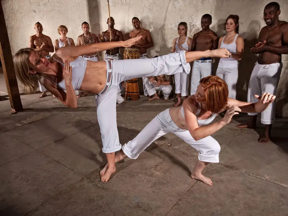 Dwie kobiety w białych strojach wykonują kopnięcia w capoeira. W tle grupa ludzi obserwuje, a jeden gra na instrumencie.