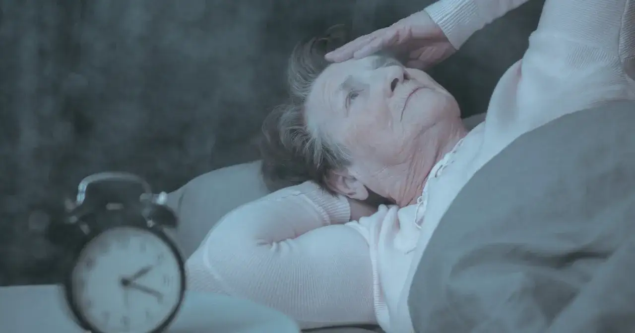 Starsza kobieta w łóżku, z ręką na czole, budzi się przy budziku. Czy to odpowiedź na pytanie, dlaczego starsi ludzie dużo śpią?
