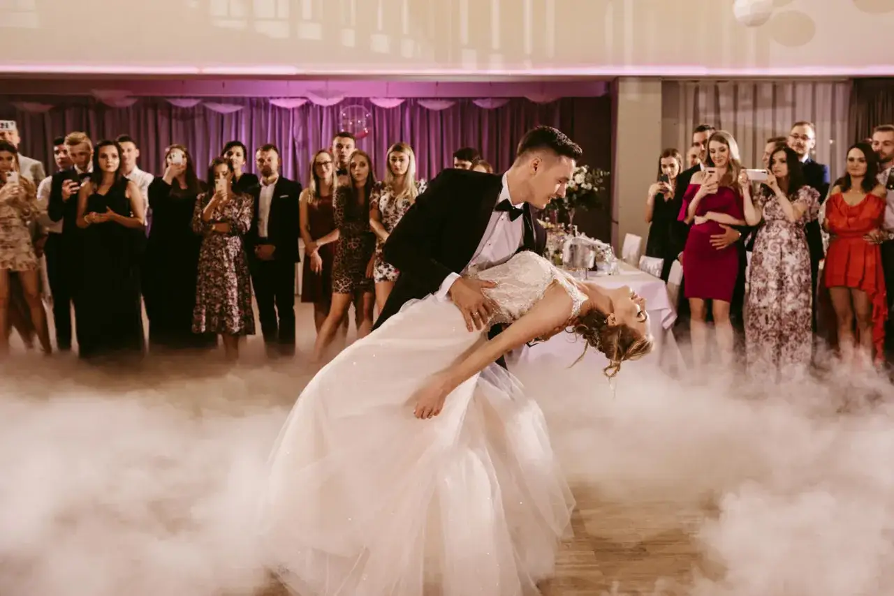 Para młoda wykonuje pierwszy taniec weselny, otoczona mgłą i gośćmi.
