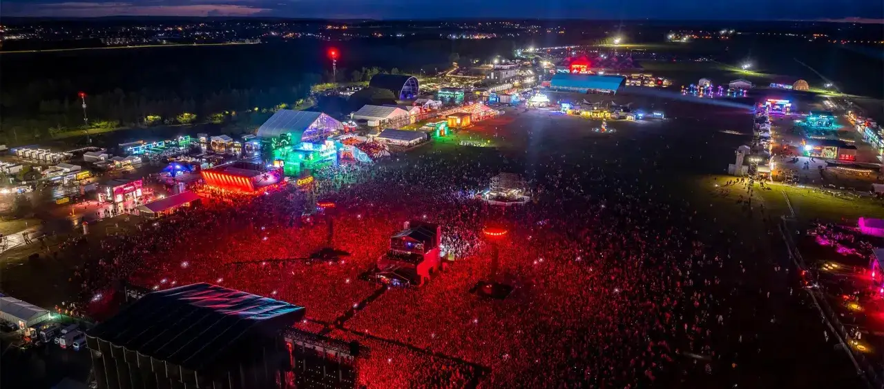 Nocny festiwal muzyczny z tłumem ludzi. Widać sceny, namioty i oświetlenie.
