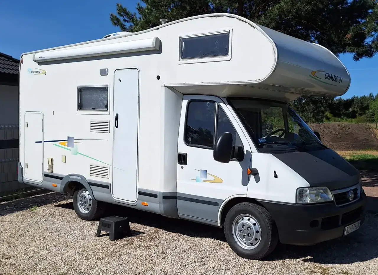 Biały kamper 4 osobowy z markizą i oknami, zaparkowany na żwirze. Idealny na rodzinne wyprawy.