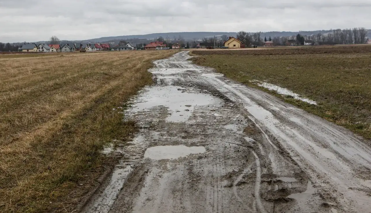 Błotnista droga dojazdowa do posesji, z licznymi kałużami i koleinami, prowadzi przez pola do zabudowań. Obowiązek gminy to zapewnienie przejezdności.