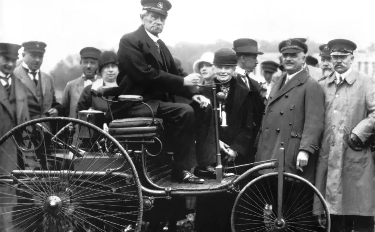Karl Benz, który wynalazł samochód, siedzi za kierownicą swojego pojazdu, otoczony przez grupę ludzi.