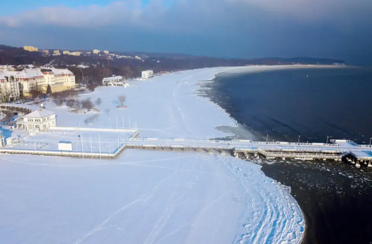 Zimowe ferie kujawsko pomorskie 2026 nad zamarzniętym morzem. Molo i plaża pokryte śniegiem, budynki w oddali.