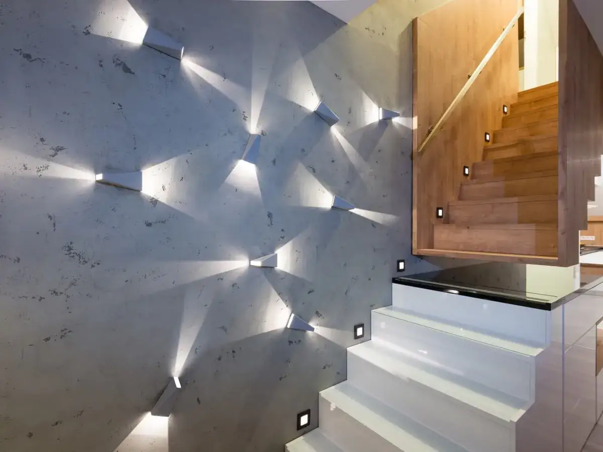 Nowoczesne schody z białymi stopniami i drewnianą balustradą. Betonowa ściana ozdobiona geometrycznymi lampami, tworzącymi ciekawe foto pod dachem.