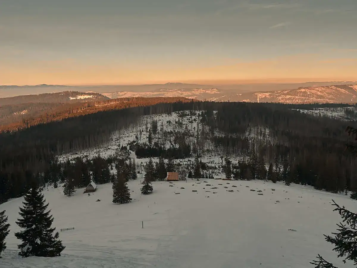Zimowy krajobraz górski o zachodzie słońca. Śnieg, lasy i małe chatki. Idealne miejsce na wędrówki, gdzie raki do chodzenia po górach zapewnią bezpieczeństwo.