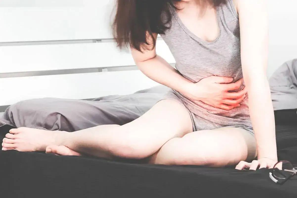 Kobieta siedzi na łóżku, trzymając się za brzuch. Czerwony obszar wskazuje ból w narządach po lewej stronie brzucha.