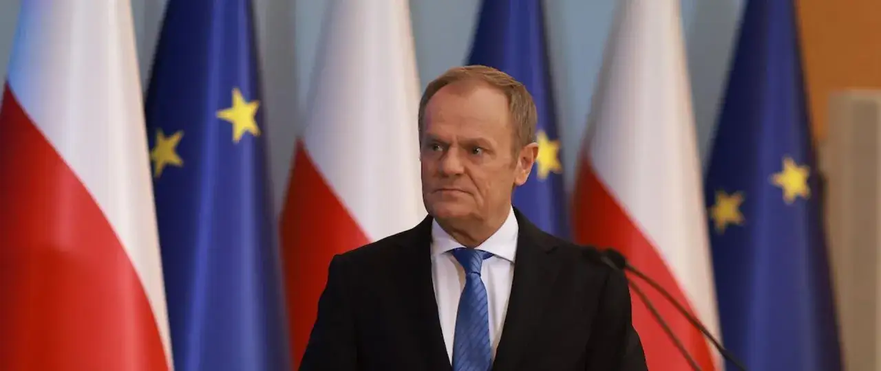 Donald Tusk w garniturze i krawacie, na tle flag Polski i Unii Europejskiej. Dziś zapewne zajmuje się ważnymi sprawami państwowymi.