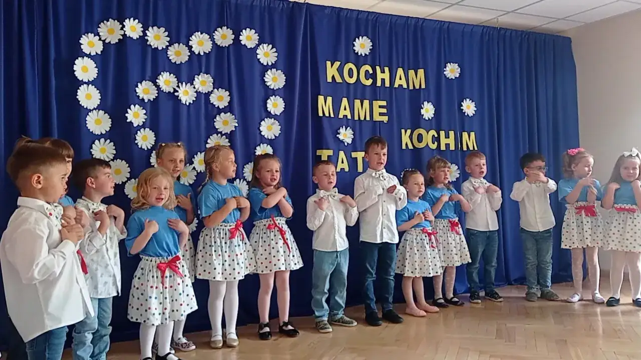 Dzieci w przedszkolu śpiewają piosenkę na dzień mamy. Na niebieskiej kurtynie ozdobionej stokrotkami widnieje napis 