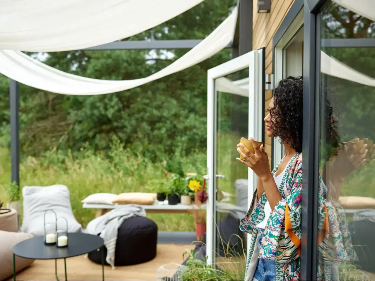 Kobieta z kubkiem kawy na małym tarasie. Inspiracja, jak urządzić mały taras: wygodne pufy, stolik i rośliny.