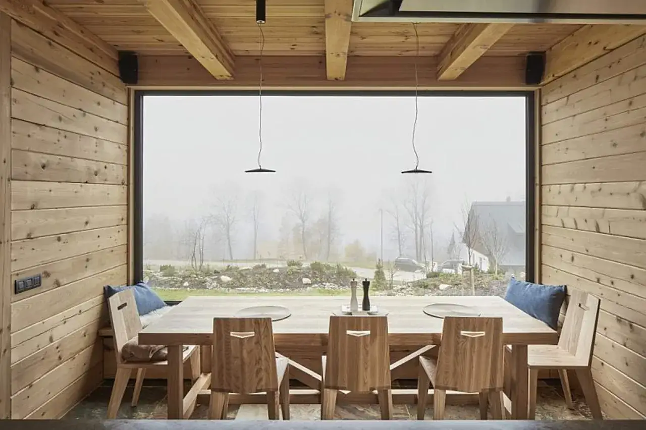 Nowoczesne domy z drewna i szkła. Drewniany st&oacute;ł i krzesła w jadalni z widokiem na mglisty krajobraz.
